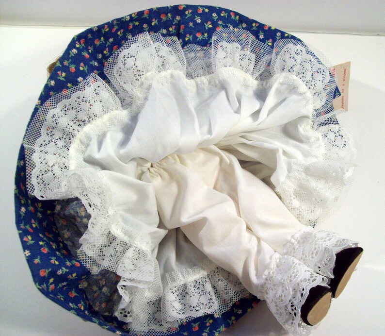 May include: A white doll with a blue and white floral print dress and white lace trim. The doll is wearing white underwear and has a white lace trim on the bottom of the dress.