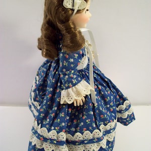 May include: A porcelain doll wearing a blue dress with white floral print and white lace trim. The doll has brown hair and is wearing a white lace headband.