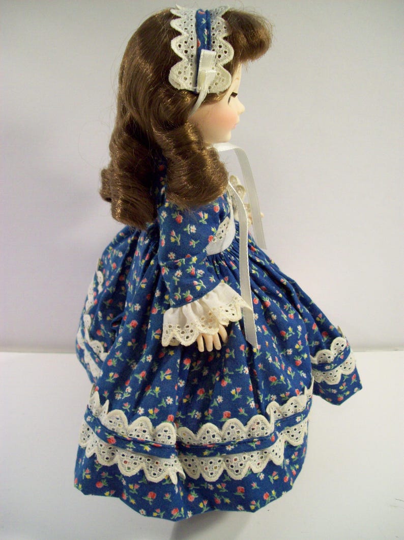 May include: A porcelain doll wearing a blue dress with white floral print and white lace trim. The doll has brown hair and is wearing a white lace headband.