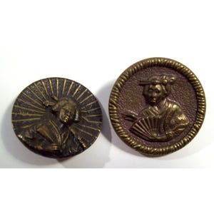 May include: Two round metal buttons with a brass finish. The left button features a figure with a sunburst background. The right button displays a figure holding a handheld fan, surrounded by a textured border with a rope-like edge design.