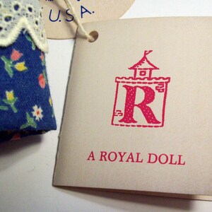 May include: A white tag with red text that reads "A Royal Doll". The tag has a red illustration of a castle with a red "R" inside.