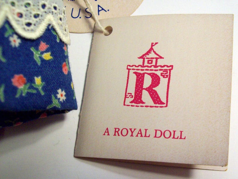 May include: A white tag with red text that reads "A Royal Doll". The tag has a red illustration of a castle with a red "R" inside.