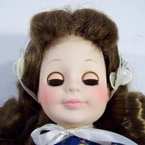 May include: A porcelain doll with brown hair styled in a bun, wearing a blue and white dress with a white bow. The doll has closed eyes with black eyelashes and pink blush on her cheeks.