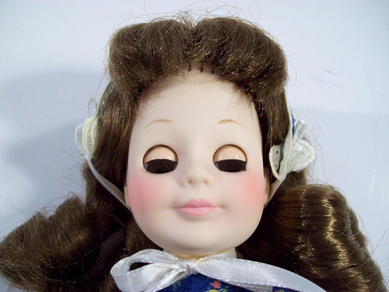 May include: A porcelain doll with brown hair styled in a bun, wearing a blue and white dress with a white bow. The doll has closed eyes with black eyelashes and pink blush on her cheeks.