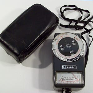 May include: A black and silver light meter with a black leather case. The light meter has a circular dial with markings for speed, ASA, and EV. The dial also has a small window that displays the meter reading. The light meter has a black and silver body with a black strap.