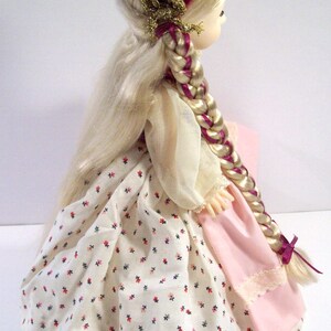 May include: A porcelain doll with long blonde hair styled in a braid. She is wearing a white dress with a floral pattern, a pink skirt, and a burgundy hat with gold trim.