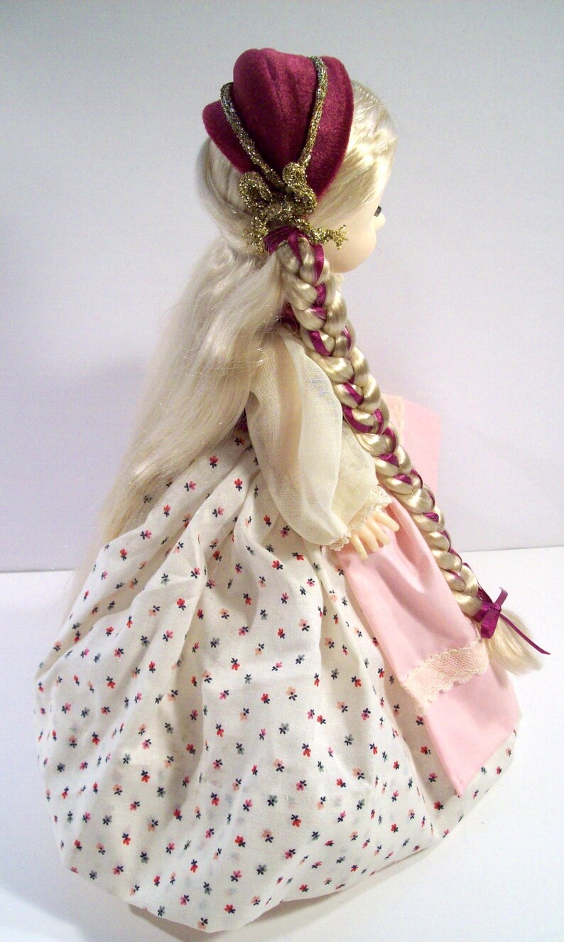 May include: A porcelain doll with long blonde hair styled in a braid. She is wearing a white dress with a floral pattern, a pink skirt, and a burgundy hat with gold trim.