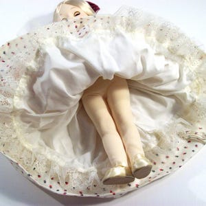 May include: A white doll with a red and white dress and a white lace trim. The doll is wearing a red hat and has blonde hair. The doll is sitting on a white surface.