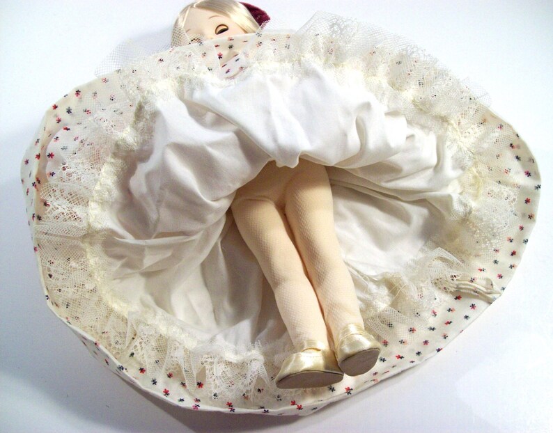 May include: A white doll with a red and white dress and a white lace trim. The doll is wearing a red hat and has blonde hair. The doll is sitting on a white surface.