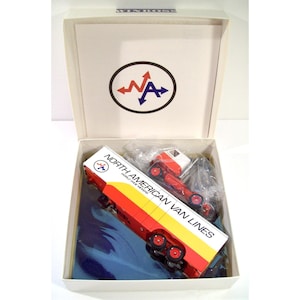 May include: A Winross die-cast model truck featuring a red and white cab and a trailer with North American Van Lines branding. The trailer displays the text North American Van Lines World Wide Movers and a logo. The model is inside an open white box.