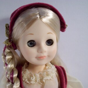 May include: A porcelain doll with blonde hair styled in braids, wearing a burgundy velvet headband and a white lace collar.
