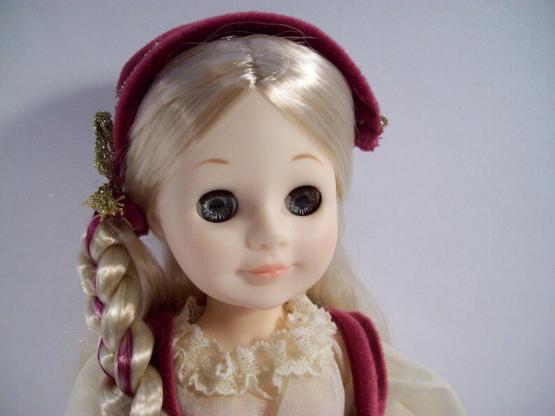 May include: A porcelain doll with blonde hair styled in braids, wearing a burgundy velvet headband and a white lace collar.