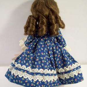 May include: A porcelain doll wearing a blue dress with a white floral pattern and white lace trim. The doll has long brown hair styled in curls.
