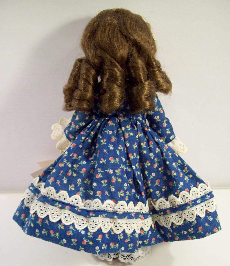 May include: A porcelain doll wearing a blue dress with a white floral pattern and white lace trim. The doll has long brown hair styled in curls.