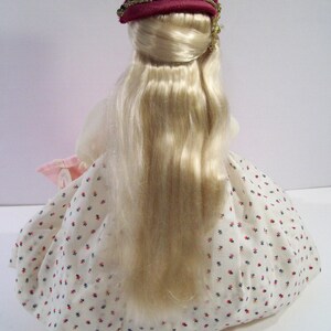 May include: A porcelain doll with long blonde hair, wearing a white dress with red and blue polka dots and a burgundy velvet hat with gold trim.