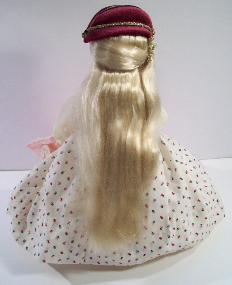 May include: A porcelain doll with long blonde hair, wearing a white dress with red and blue polka dots and a burgundy velvet hat with gold trim.