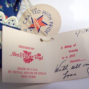 May include: A white tag with red text that reads "Designs by Miss Elsa go Royal" and "Made in U.S.A. by Royal House of Dolls New York". The tag is attached to a larger white tag with red and blue text that reads "A thing of beauty is a JOY forever... With all my love Elsa".