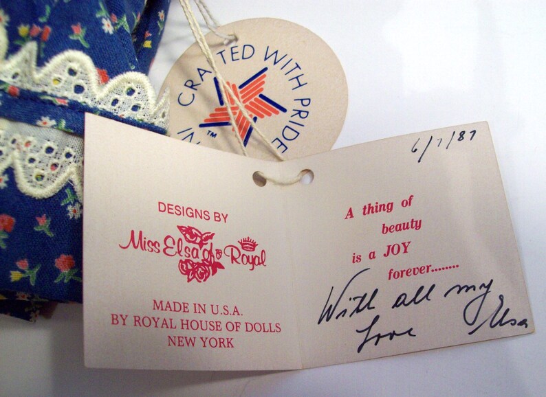 May include: A white tag with red text that reads "Designs by Miss Elsa go Royal" and "Made in U.S.A. by Royal House of Dolls New York". The tag is attached to a larger white tag with red and blue text that reads "A thing of beauty is a JOY forever... With all my love Elsa".