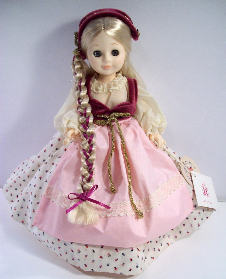 May include: A porcelain doll wearing a pink dress with white polka dots, a burgundy velvet vest, and a burgundy velvet hat. The doll has blonde hair in a braid with a burgundy ribbon. The doll is holding a tag that says "A Royal Doll".