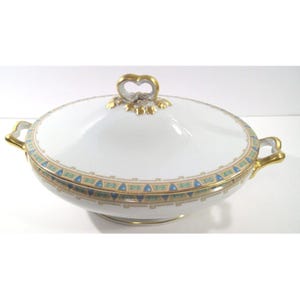 May include: White porcelain serving dish with a lid, featuring gold accents and a decorative border with blue and tan geometric patterns. The dish has a heart-shaped handle on the lid and gold handles on each side.