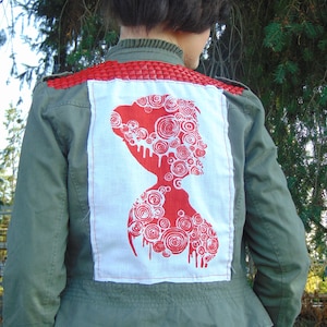 May include: An olive green jacket with a red woven shoulder detail and a large white patch on the back. The patch features a red silhouette design with spiral and drip details. The jacket is worn with blue jeans.