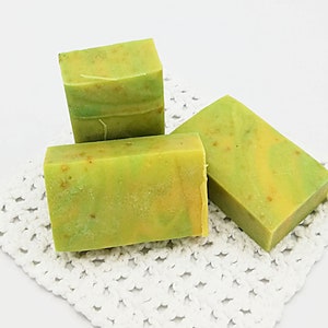 以下が含まれることがあります： 緑と黄色の渦巻き模様のハンドメイド石鹸が3つ。石鹸は白いレースのドイリーの上に置かれています。