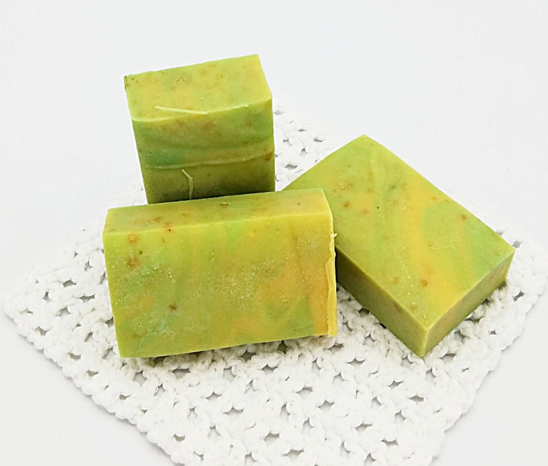 以下が含まれることがあります： 緑と黄色の渦巻き模様のハンドメイド石鹸が3つ。石鹸は白いレースのドイリーの上に置かれています。