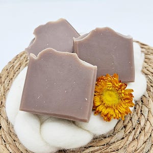 Puede incluir: Tres barras de jabón artesanal de color morado claro, dispuestas en una cesta tejida con una superficie blanca y esponjosa y una flor amarilla.