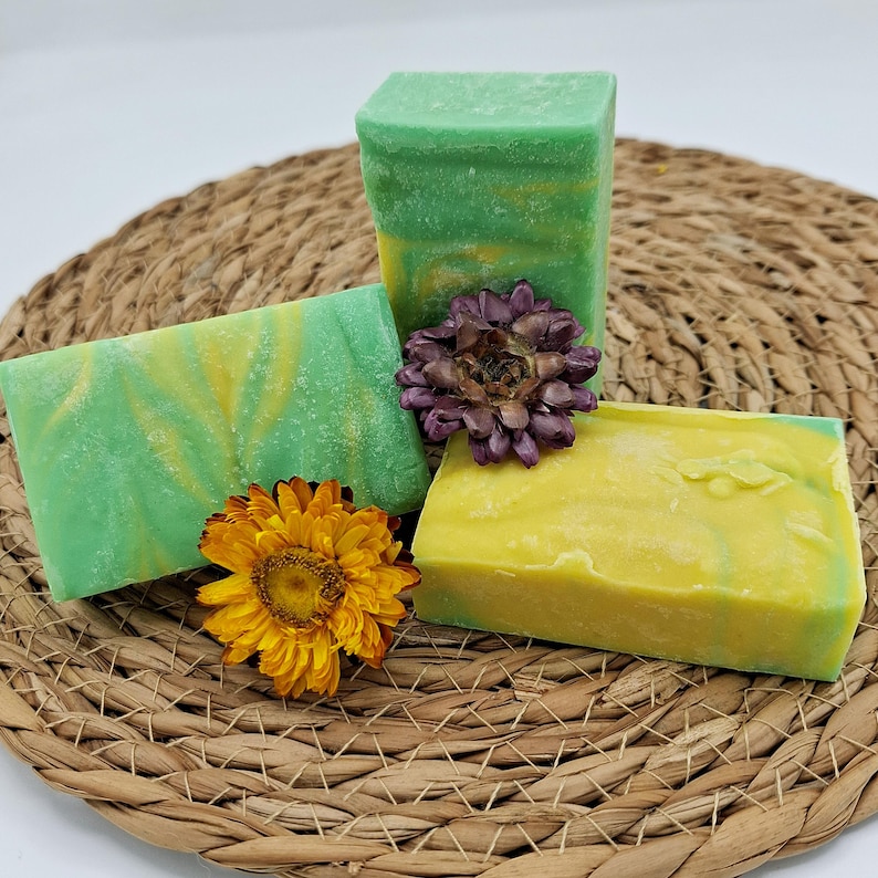 以下が含まれることがあります： 手作りの石鹸が3つ、緑と黄色の渦巻き模様、黄色と緑色の渦巻き模様、無地の緑色の石鹸。石鹸は、茶色の籐製のマットの上に置かれ、2つのドライフラワーが添えられています。