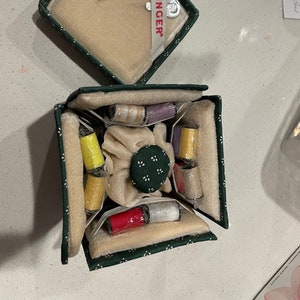 May include: A green fabric sewing box with a white floral pattern. The box is open and contains a variety of spools of thread in different colors, including yellow, red, white, and purple. The box also has a small, round, green button with white dots.