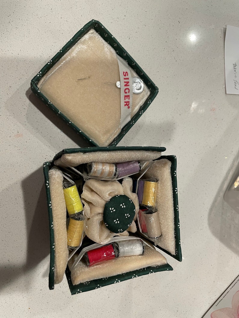 May include: A green fabric sewing box with a white floral pattern. The box is open and contains a variety of spools of thread in different colors, including yellow, red, white, and purple. The box also has a small, round, green button with white dots.