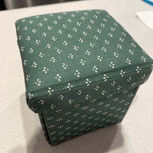 May include: A green fabric storage box with a white floral pattern. The box is square and has a hinged lid.