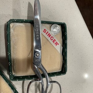 May include: A pair of silver sewing scissors with a green and white box. The box has a white ribbon with the word "SINGER" printed on it. The scissors have the word "SINGER" and the number "609" printed on them.