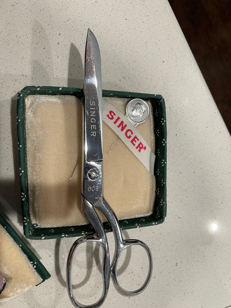 May include: A pair of silver sewing scissors with a green and white box. The box has a white ribbon with the word "SINGER" printed on it. The scissors have the word "SINGER" and the number "609" printed on them.