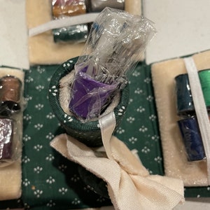 May include: A green and white fabric sewing kit with a purple container of needles, spools of thread, and a white fabric bow.