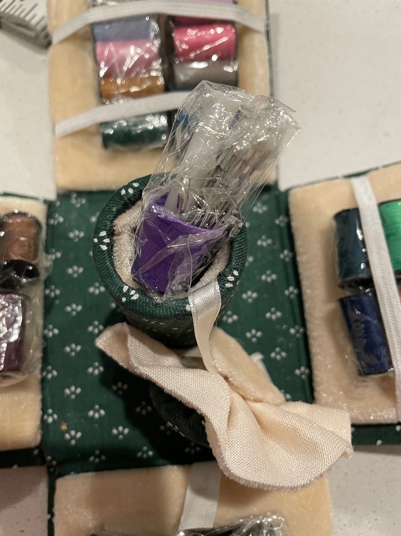 May include: A green and white fabric sewing kit with a purple container of needles, spools of thread, and a white fabric bow.