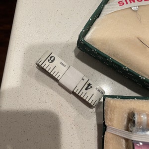 May include: A white sewing tape measure with black markings, reading "60 inch" and "Made in China". The tape measure is lying on a white countertop. A green and white box with the word "SINGER" in red letters is in the background.