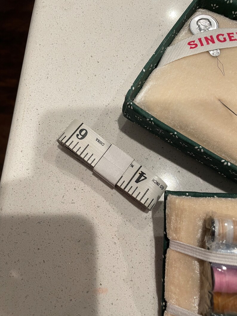 May include: A white sewing tape measure with black markings, reading "60 inch" and "Made in China". The tape measure is lying on a white countertop. A green and white box with the word "SINGER" in red letters is in the background.