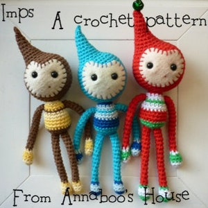 Puede incluir: Tres duendes de crochet, uno marrón, uno azul y uno rojo, con detalles verdes, blancos y amarillos. Los duendes tienen ojos grandes y sombreros puntiagudos. Los duendes están de pie sobre una superficie blanca. "Imps A crochet pattern from Annaboo's House" está escrito debajo de los duendes.