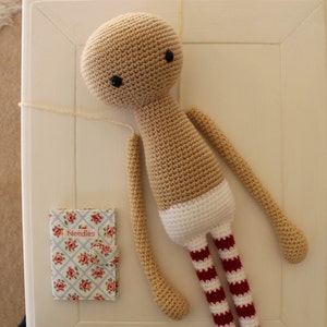 May include: A crocheted doll with a beige body, white diaper, and red and white striped legs. The doll has black eyes and is lying on a white surface.
