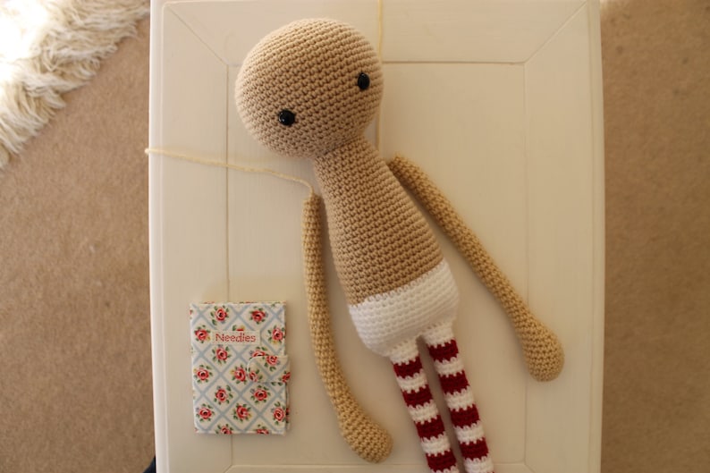 May include: A crocheted doll with a beige body, white diaper, and red and white striped legs. The doll has black eyes and is lying on a white surface.
