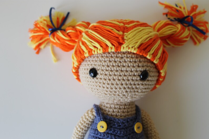 May include: A crocheted doll with orange and yellow yarn hair, brown eyes, and a blue denim overall dress with yellow buttons.