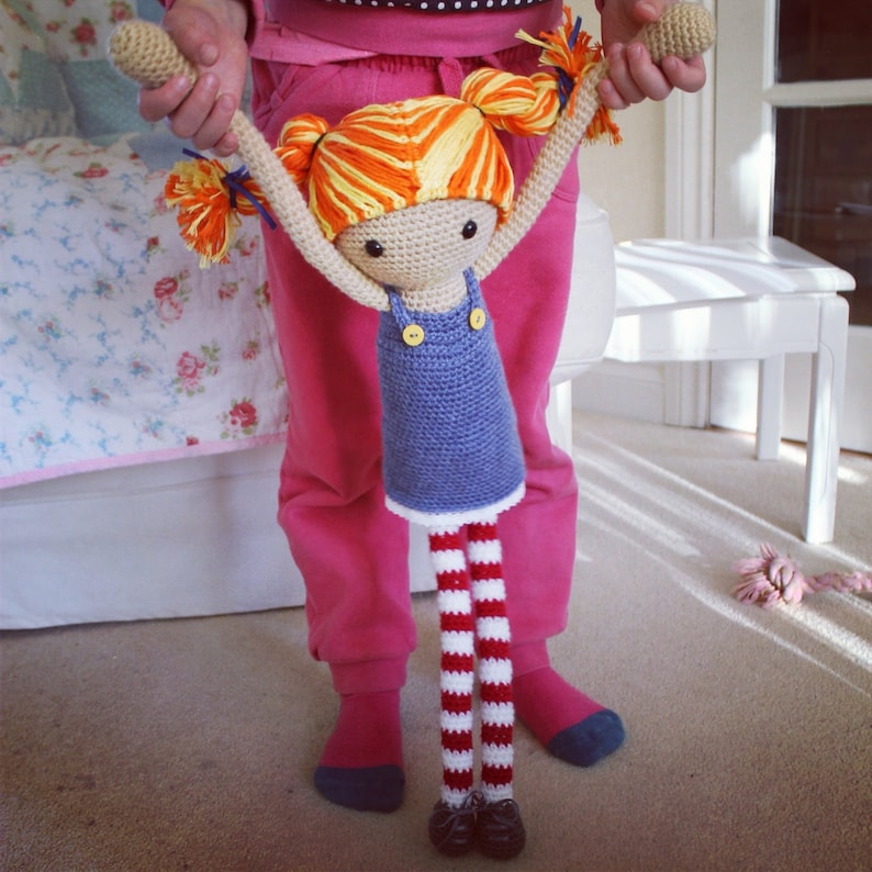 May include: A crocheted doll with orange and yellow hair, a blue dress, and red and white striped legs. The doll is being held by a person wearing pink pants.
