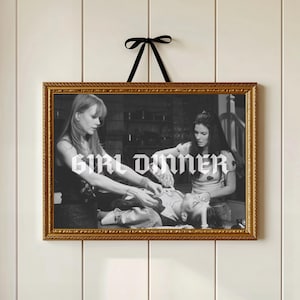 May include: A framed black and white print with the text "GIRL DINNER" in a gothic font. The gold frame is rectangular and has ornate detailing. A black ribbon is attached to the top for hanging.
