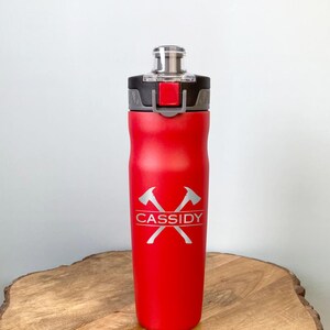 May include: A red water bottle with a black and silver lid. The bottle has a silver graphic of crossed axes and the name "Cassidy". The bottle is cylindrical and sits on a wooden slab.