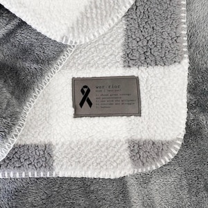 May include: A gray and white checkered fleece blanket with a tag that reads "warrior" and includes a ribbon. The tag has a message about showing great courage and perseverance.