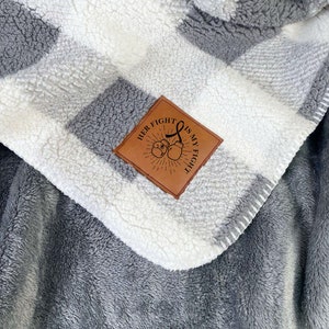 May include: A gray and white checkered fleece blanket with a brown leather patch that says "Her Fight Is My Fight" with a pair of boxing gloves and a sunburst design.