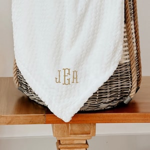 May include: A white plush throw blanket with a monogrammed "J.E.M." in gold thread, folded and placed in a wicker basket with rope handles.