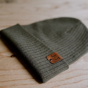 May include: A dark green knitted beanie with a brown leather patch that says "JT" in white lettering.