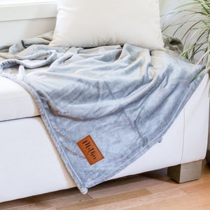 May include: A soft, gray throw blanket with a brown leather tag that says "Mellow" is draped over a white couch. A white pillow is on the couch behind the blanket. A potted plant with green leaves is in the background.